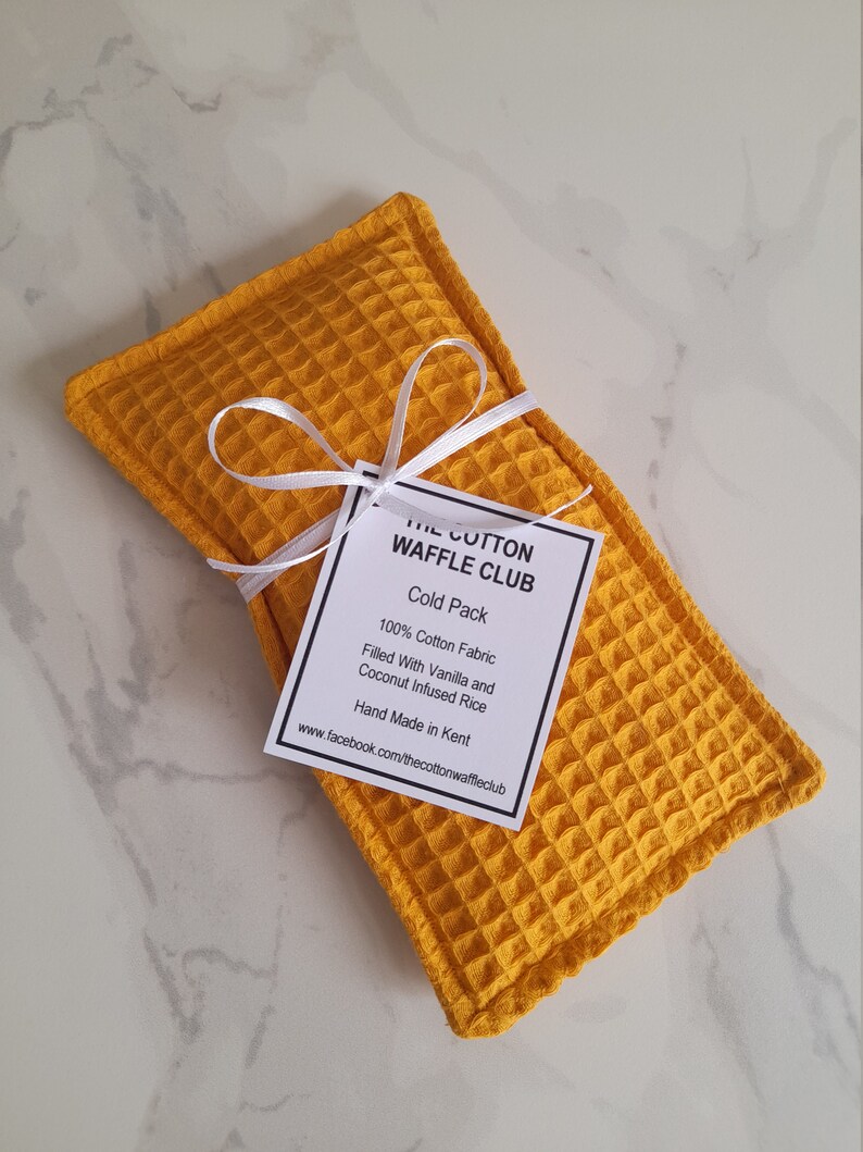 May include: A rectangular mustard yellow cold pack made of waffle-textured cotton fabric. A white ribbon is tied around the center, holding a white tag that reads: THE COTTON WAFFLE CLUB, Cold Pack, 100% Cotton Fabric, Filled With Vanilla and Coconut Infused Rice, Hand Made in Kent, www.facebook.com/thecottonwaffleclub.