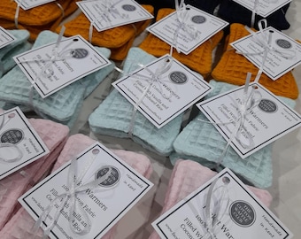 May include: A collection of square pocket warmers in various colors including pink, light blue, orange, and dark blue. Each warmer features a waffle-textured cotton fabric, tied with a white ribbon and a tag labeled Pocket Warmers, 100% Cotton Fabric.