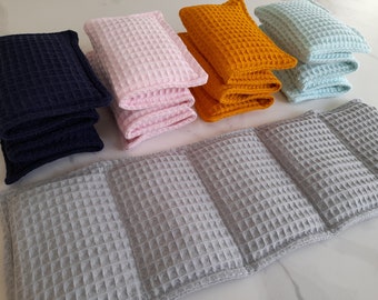 May include: A collection of rectangular waffle-weave fabric items in navy blue, light pink, mustard yellow, and pale blue, arranged in stacks, with a long, segmented gray waffle-weave fabric piece positioned horizontally in the foreground.