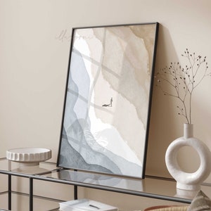 Puede incluir: Una pintura abstracta de acuarela enmarcada con un diseño minimalista que presenta tonos de azul, gris y beige. Una pequeña figura se representa caminando por un sendero en el centro de la pintura.