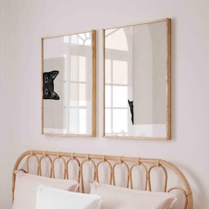 May include: Two framed prints of black cats peeking out from behind a white wall. The prints are hung on a light pink wall above a wicker headboard.