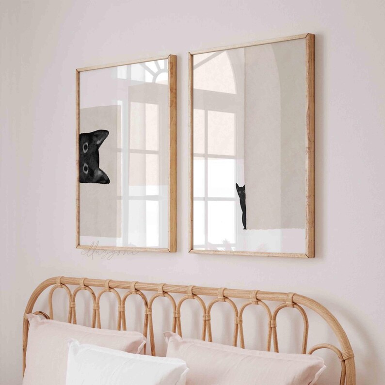 May include: Two framed prints of black cats peeking out from behind a white wall. The prints are hung on a light pink wall above a wicker headboard.