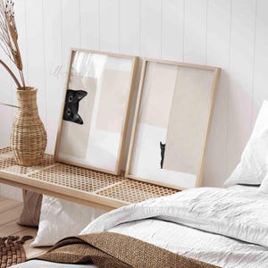 May include: Two framed prints with black and white images of a cat peeking out from behind a beige background. The frames are light wood and are hanging on a white wall.