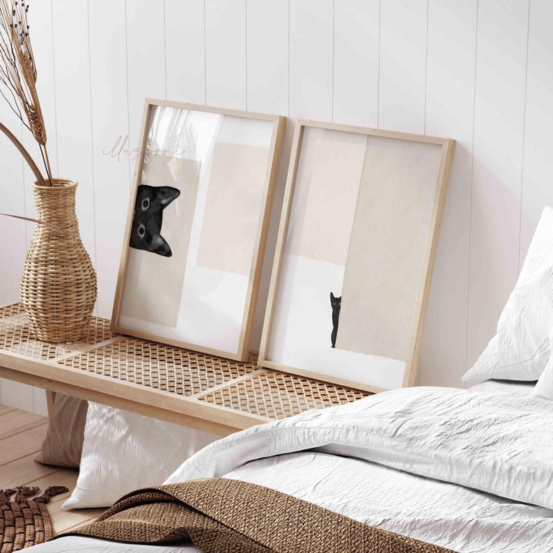 May include: Two framed prints with black and white images of a cat peeking out from behind a beige background. The frames are light wood and are hanging on a white wall.