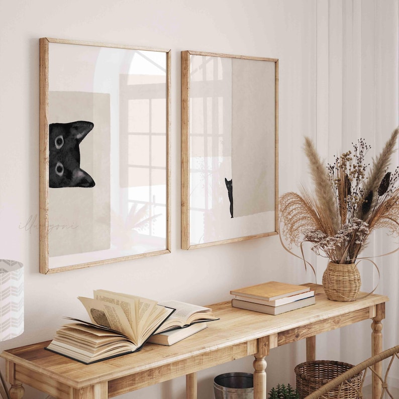 May include: Two framed black and white illustrations of a black cat. The cat in the left frame is looking up at the viewer, while the cat in the right frame is walking away. The frames are wooden and have a light brown finish.