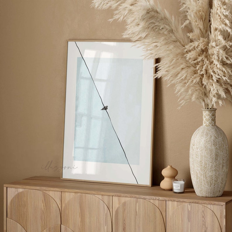 May include: A framed print with a light blue background and a single black bird silhouette on a diagonal line. The print is hanging on a light wood cabinet with a vase of dried pampas grass.