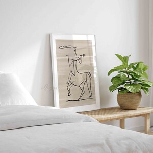 Op de afbeelding: Een zwart-witte lijntekening van een centaur die een muziekinstrument bespeelt. De tekening is op een beige achtergrond en ingelijst in een wit frame. De tekening hangt aan een witte muur boven een wit bed met een wit hoofdeinde.