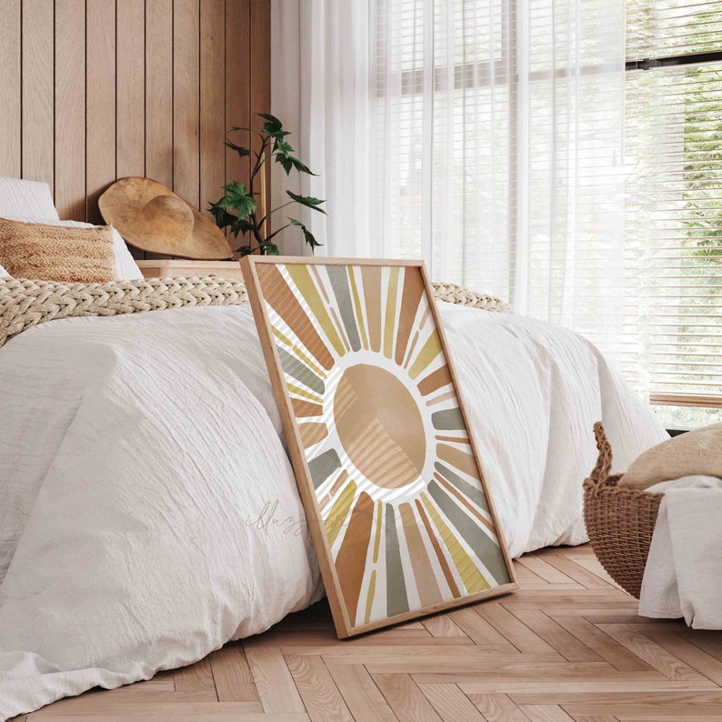 May include: A framed print of a stylized sun with rays in shades of brown, yellow, and green. The sun is set against a white background. The print is leaning against a white bed with a wooden headboard.