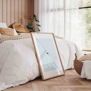 May include: A framed print of a minimalist landscape with a single bird perched on a wire against a light blue sky. The print is leaning against a white bed with a wooden headboard and a woven throw blanket.