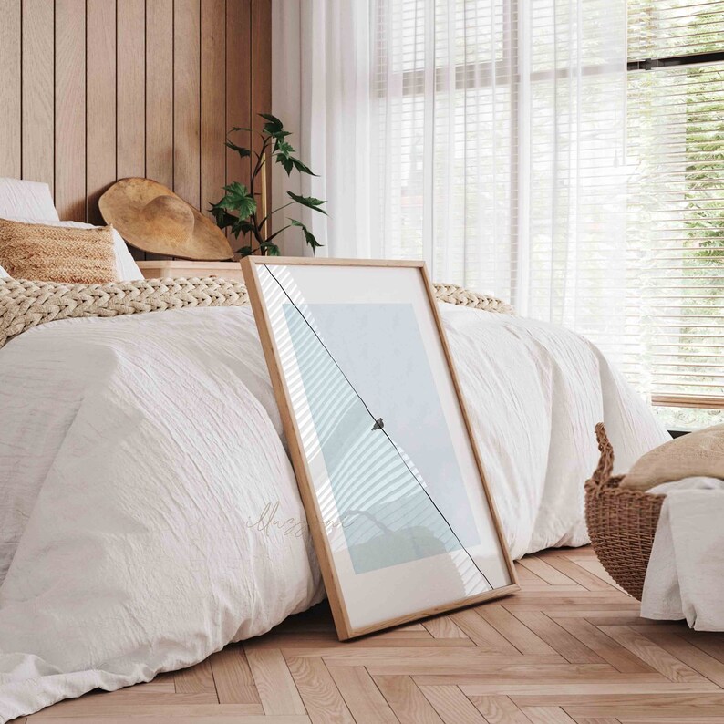 May include: A framed print of a minimalist landscape with a single bird perched on a wire against a light blue sky. The print is leaning against a white bed with a wooden headboard and a woven throw blanket.