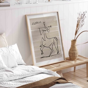 Op de afbeelding: Een ingelijst afdruk van een zwart-witte lijntekening van een persoon die op een paard rijdt. De tekening is op een beige achtergrond. De afdruk hangt aan een witte muur boven een houten bank.