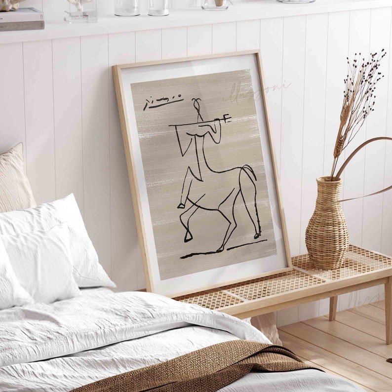 Op de afbeelding: Een ingelijst afdruk van een zwart-witte lijntekening van een persoon die op een paard rijdt. De tekening is op een beige achtergrond. De afdruk hangt aan een witte muur boven een houten bank.