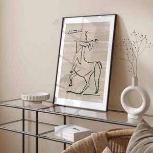 Op de afbeelding: Een zwart-witte lijntekening van een centaur, ingelijst in een zwart frame, hangt aan een muur boven een glazen plank. De tekening is op een beige achtergrond.
