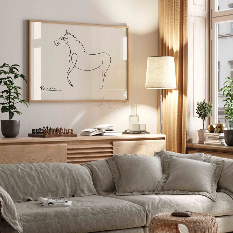 May include: A framed black and white line drawing of a horse, with the artist's signature "Picasso" in the bottom left corner. The drawing is hanging on a white wall above a wooden shelf with a chessboard and books.