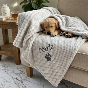 Pode incluir: Uma manta cinza-clara com textura de tecido waffle, apresentando o nome Narla bordado em letras cursivas cinza-escuras acima de um &iacute;cone de pata cinza-escuro, drapeada sobre um filhote de golden retriever dormindo em um sof&aacute;.