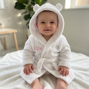 Peut inclure: Un peignoir blanc à capuche avec des oreilles d'ours, une ceinture à nouer, le prénom Ellie brodé en rose sur la poitrine et un petit motif d'éléphant gris brodé sur le côté opposé de la poitrine.