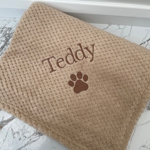 Pode incluir: Uma manta bege com textura de tecido waffle, apresentando o nome Teddy bordado em fio castanho escuro, acompanhado por um &iacute;cone de pata bordado em castanho escuro centrado abaixo do nome no tecido.