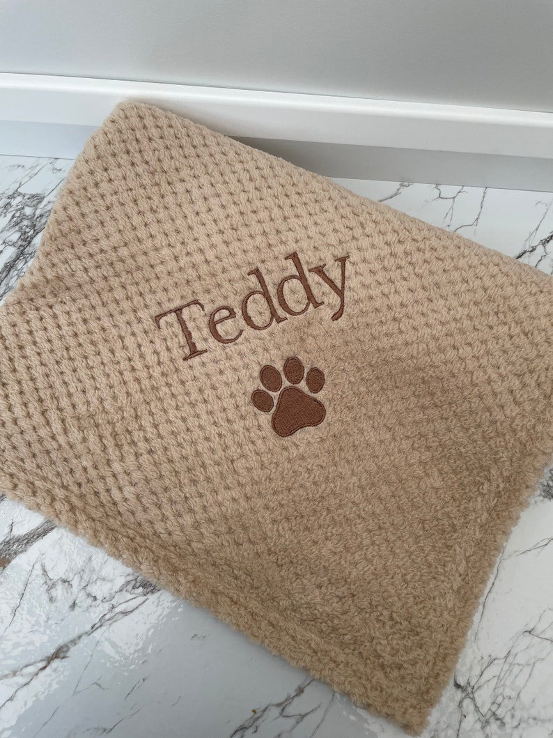 Pode incluir: Uma manta bege com textura de tecido waffle, apresentando o nome Teddy bordado em fio castanho escuro, acompanhado por um &iacute;cone de pata bordado em castanho escuro centrado abaixo do nome no tecido.