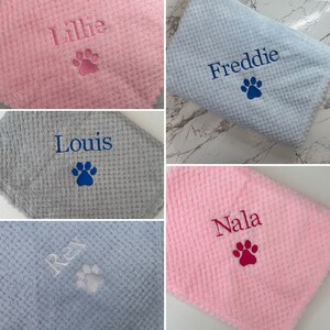 Pode incluir: Uma cole&ccedil;&atilde;o de cinco mantas macias com textura de waffle em rosa, azul claro e cinza. Cada manta apresenta bordados personalizados com os nomes Lillie, Freddie, Louis, Rex e Nala, acompanhados por um &iacute;cone de pata bordado.