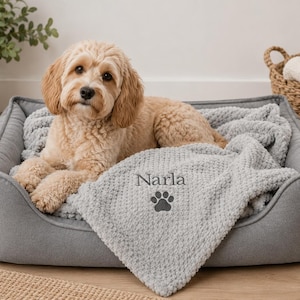 Pode incluir: Um c&atilde;o de pelo claro e ondulado descansa numa cama retangular cinzenta para animais. Uma manta cinzenta texturizada est&aacute; sobre a cama, apresentando o nome Narla bordado acima de um &iacute;cone de pata cinzento-escuro centrado.