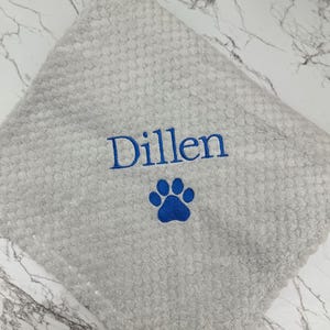 Pode incluir: Um pano quadrado de cor cinza-claro com textura, apresentando o nome Dillen bordado em fio azul sobre um desenho de pegada de pata tamb&eacute;m bordado em azul. O tecido tem uma superf&iacute;cie macia com uma textura de favo de mel.