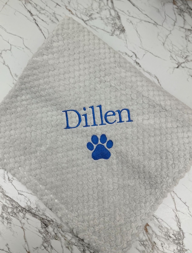 Pode incluir: Um pano quadrado de cor cinza-claro com textura, apresentando o nome Dillen bordado em fio azul sobre um desenho de pegada de pata tamb&eacute;m bordado em azul. O tecido tem uma superf&iacute;cie macia com uma textura de favo de mel.