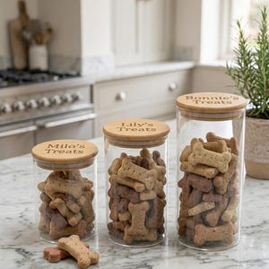 Pode incluir: Tr&ecirc;s potes cil&iacute;ndricos de vidro transparente com tampas de madeira clara, cheios de petiscos para c&atilde;es em forma de osso. As tampas est&atilde;o gravadas com os nomes Milo's Treats, Lily's Treats e Bonnie's Treats.