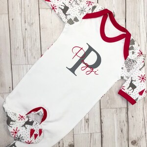 May include: White baby onesie with red trim and long sleeves. The sleeves and feet have a gray and white pattern with red snowflakes, trees, and reindeer. The onesie has a gray letter "P" with the name "Poppy" written in red underneath.