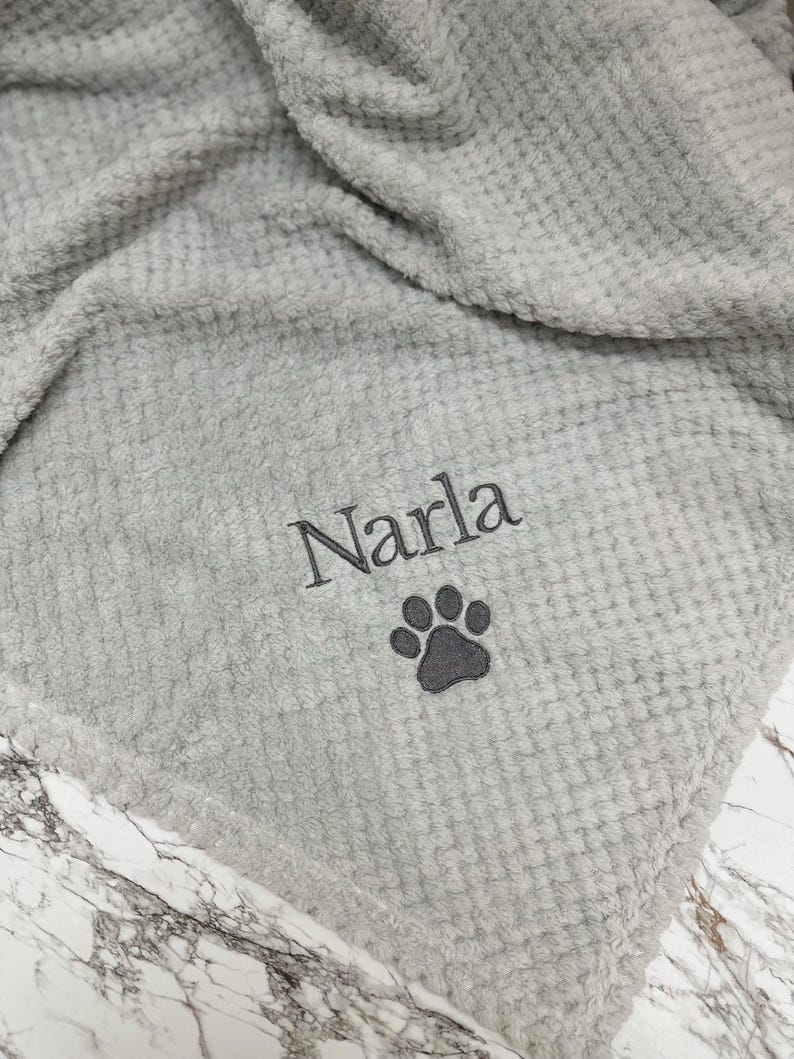 Pode incluir: Uma manta cinza-claro com textura de waffle apresentando o nome bordado Narla em fio cinza-escuro, posicionado acima de um &iacute;cone de pata bordado em cinza-escuro. O tecido tem uma apar&ecirc;ncia macia e felpuda com um padr&atilde;o de grade.