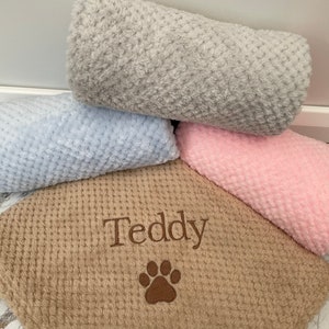 Pode incluir: Uma cole&ccedil;&atilde;o de quatro cobertores macios e texturizados em cinza, azul claro, rosa e bege. O cobertor bege apresenta o nome Teddy bordado em fio marrom acima de um desenho de pata bordado em marrom, com um padr&atilde;o de tecido tipo waffle.