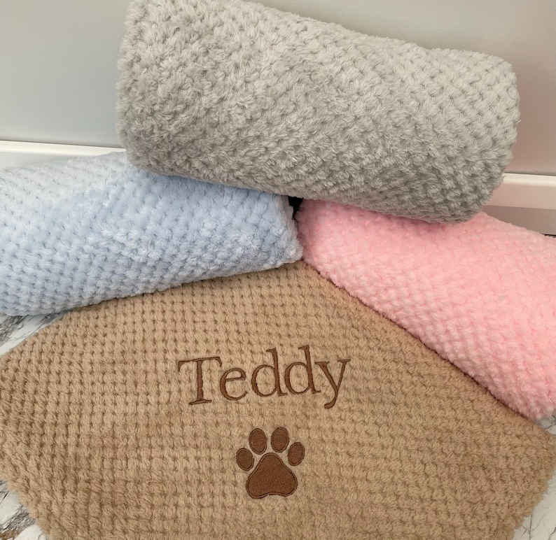 Pode incluir: Uma cole&ccedil;&atilde;o de quatro cobertores macios e texturizados em cinza, azul claro, rosa e bege. O cobertor bege apresenta o nome Teddy bordado em fio marrom acima de um desenho de pata bordado em marrom, com um padr&atilde;o de tecido tipo waffle.