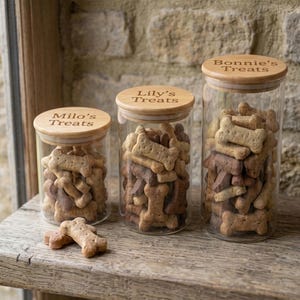 May include: Three clear glass cylindrical storage jars with light wood lids, each filled with bone-shaped dog treats. The lids are engraved with the text Milo's Treats, Lily's Treats, and Bonnie's Treats. One treat sits outside the jars.