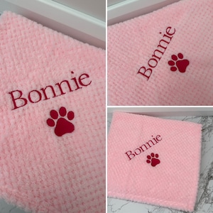 Pode incluir: Um quadrado de tecido texturizado rosa claro macio com o nome Bonnie bordado em fio rosa escuro, acompanhado por um design de pata bordado em rosa escuro posicionado diretamente abaixo do nome na superf&iacute;cie do tecido.