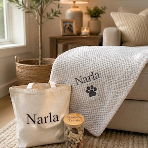 Pode incluir: Uma manta de malha waffle cor de creme com o nome Narla bordado e uma pata cinzento-escura, um saco de lona creme a condizer com o nome Narla impresso em castanho escuro e um frasco de vidro com tampa de madeira e Narla's Treats.