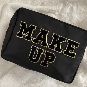 以下が含まれることがあります： 大きなバーシティスタイルの文字で「MAKE UP」と書かれた長方形の黒いコスメポーチ。文字は黒色で、ゴールドのグリッターの縁取りと質感のあるふわふわとした仕上げが施されており、滑らかな黒い生地のポーチの前面中央に配置されています。