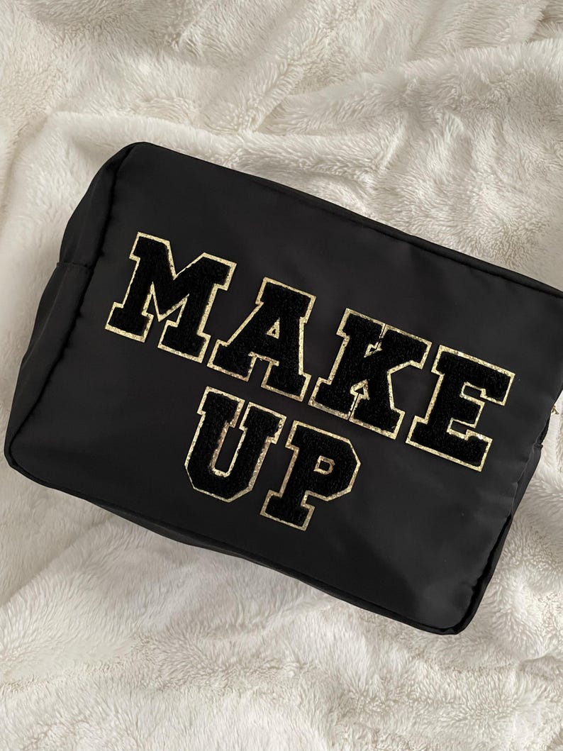 以下が含まれることがあります： 大きなバーシティスタイルの文字で「MAKE UP」と書かれた長方形の黒いコスメポーチ。文字は黒色で、ゴールドのグリッターの縁取りと質感のあるふわふわとした仕上げが施されており、滑らかな黒い生地のポーチの前面中央に配置されています。