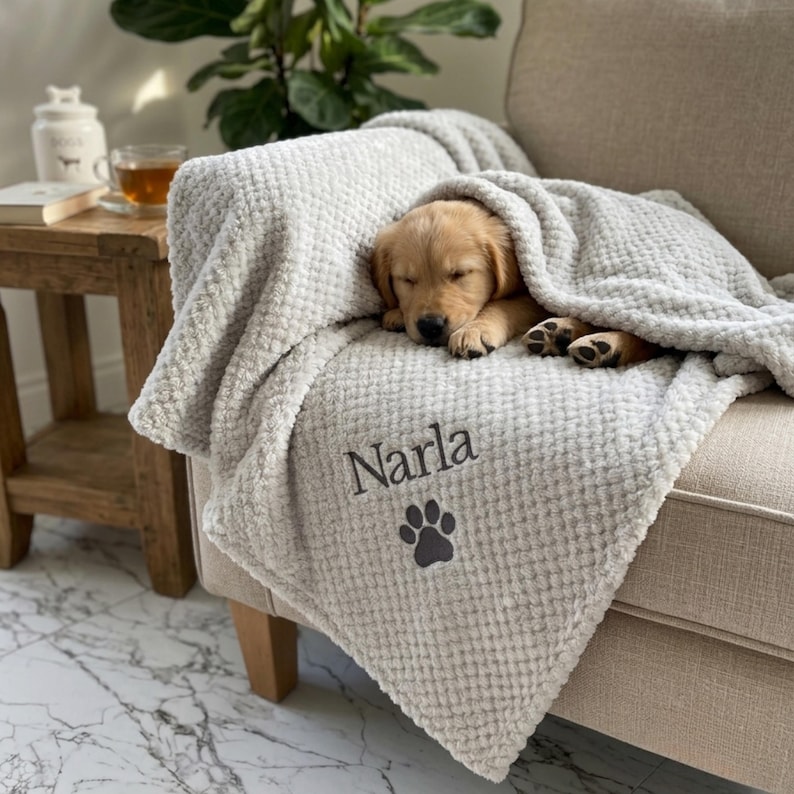 Pode incluir: Uma manta cinza-clara com textura de tecido waffle, apresentando o nome Narla bordado em letras cursivas cinza-escuras acima de um &iacute;cone de pata cinza-escuro, drapeada sobre um filhote de golden retriever dormindo em um sof&aacute;.