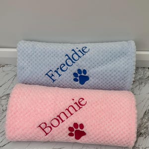 Pode incluir: Duas mantas enroladas com textura em relevo, uma azul-clara e uma rosa-clara. A manta azul est&aacute; bordada com o nome Freddie em fio azul-escuro ao lado de uma pata. A manta rosa apresenta o nome Bonnie em fio rosa-escuro com uma pata.