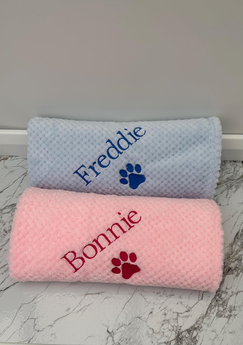 Pode incluir: Duas mantas enroladas com textura em relevo, uma azul-clara e uma rosa-clara. A manta azul est&aacute; bordada com o nome Freddie em fio azul-escuro ao lado de uma pata. A manta rosa apresenta o nome Bonnie em fio rosa-escuro com uma pata.