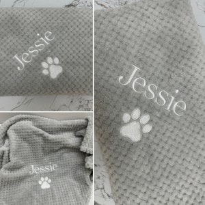 Pode incluir: Uma manta cinzenta macia com textura de tecido waffle, apresentando um bordado branco do nome Jessie e uma pegada de pata. O tecido tem um padr&atilde;o de grelha em relevo em toda a superf&iacute;cie e o bordado est&aacute; claramente posicionado.