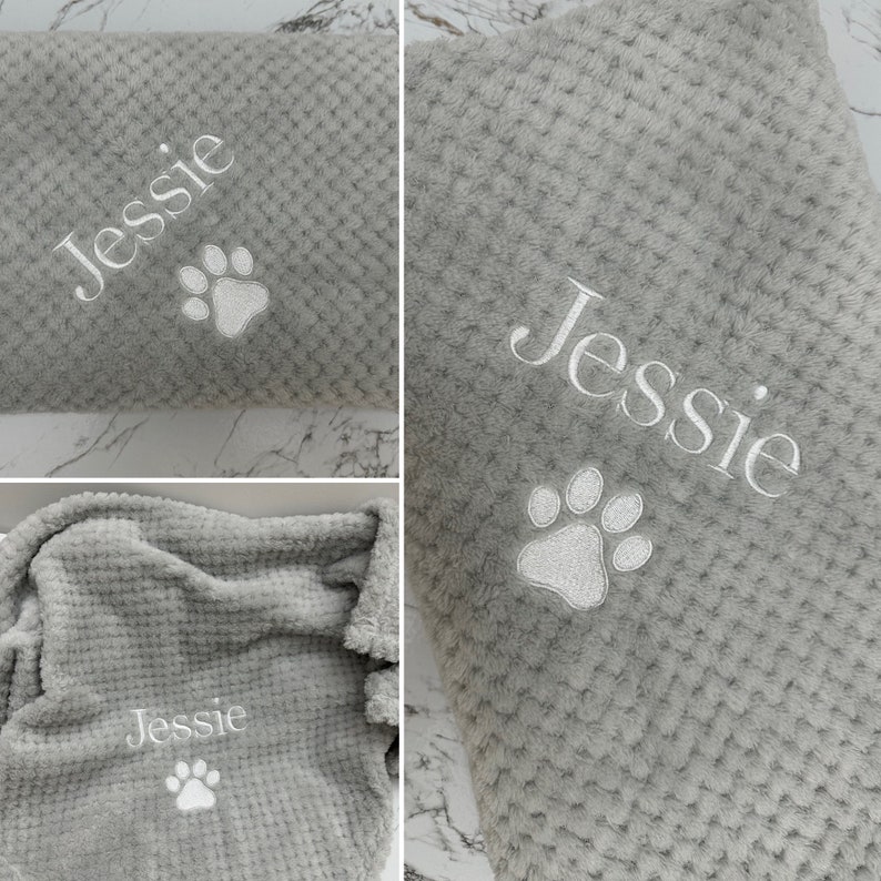 Pode incluir: Uma manta cinzenta macia com textura de tecido waffle, apresentando um bordado branco do nome Jessie e uma pegada de pata. O tecido tem um padr&atilde;o de grelha em relevo em toda a superf&iacute;cie e o bordado est&aacute; claramente posicionado.