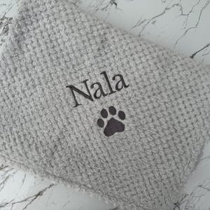Pode incluir: Uma manta cinza-claro com textura de waffle apresentando o nome Nala bordado em fio cinza-escuro, posicionado acima de um &iacute;cone de pata bordado em cinza-escuro. O tecido tem um padr&atilde;o de grade macio e elevado em toda a superf&iacute;cie.