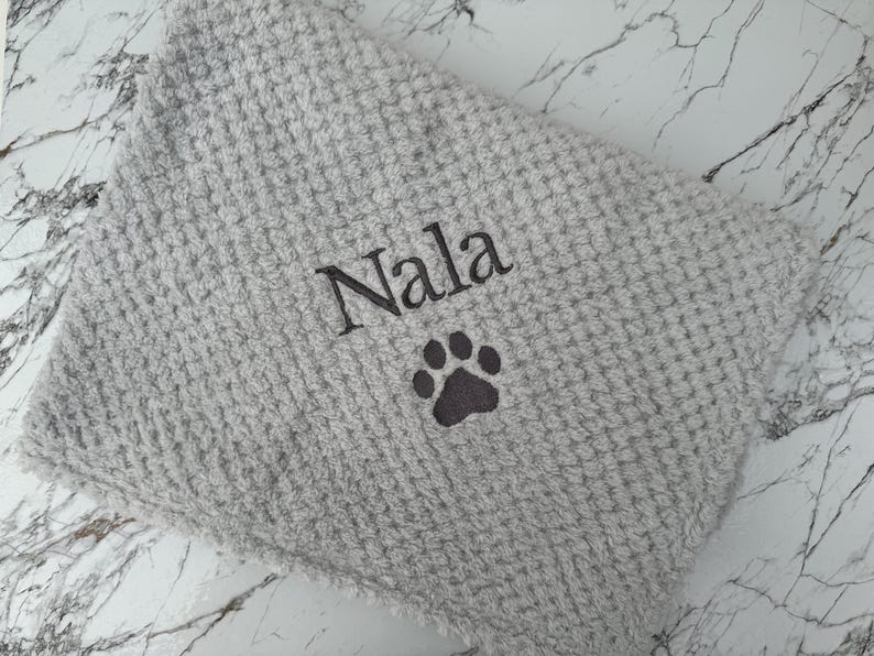Pode incluir: Uma manta cinza-claro com textura de waffle apresentando o nome Nala bordado em fio cinza-escuro, posicionado acima de um &iacute;cone de pata bordado em cinza-escuro. O tecido tem um padr&atilde;o de grade macio e elevado em toda a superf&iacute;cie.
