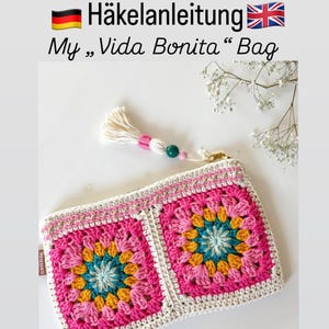 Può includere: Una borsa all'uncinetto con un colorato motivo floreale. La borsa ha una chiusura a cerniera e un nappa con un dettaglio rosa. Il testo sull'immagine recita "Häkelanleitung My Vida Bonita Bag".