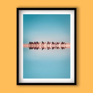 May include: A framed print featuring a line of camels with riders walking along a beach, reflected in the water below. The sky is a clear blue, and the sand is a light tan color. The image is framed in a black border.