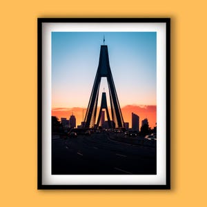 Puede incluir: Impresión enmarcada de un puente al atardecer. El puente tiene un diseño arquitectónico único con dos torres altas en forma de A. El cielo pasa del azul al naranja y al rojo, con edificios de la ciudad al fondo. Se ven coches en la carretera.