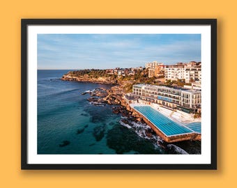 May include: A framed landscape photograph showing an aerial view of a coastal swimming pool built into rocky cliffs beside the ocean. The scene features turquoise water, a multi-story white building, and residential houses under a clear blue sky.