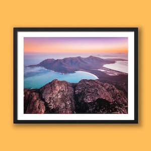 May include: A framed landscape photograph showing a mountain range, turquoise water, and a bay. The sky transitions from orange to purple. The photograph is in a black frame and is displayed against a yellow background.