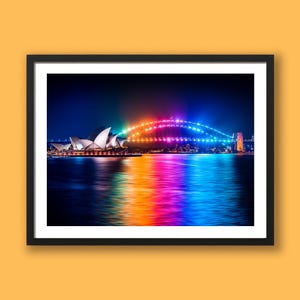 May include: A framed print depicting Sydney, Australia at night. The Sydney Opera House and Harbour Bridge are lit with vibrant colours. The water reflects the rainbow lights, creating a beautiful scene. The frame is black.
