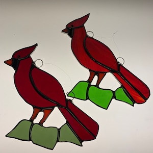 May include: Two stained glass cardinal birds perched on green leaves. The cardinals are red with black outlines and the leaves are a light green.